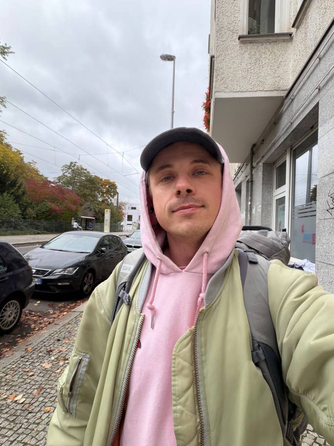 A young man in a green jacket and pink hoodie stands on a city street under cloudy skies.