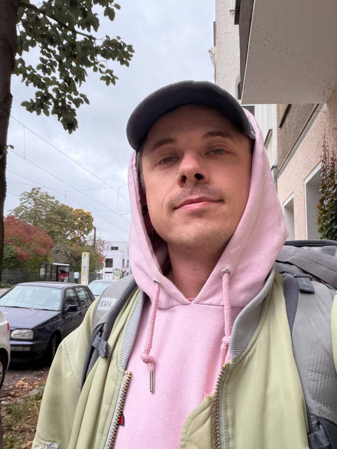 A person wearing a pink hoodie and a green jacket is standing outdoors with cars and trees