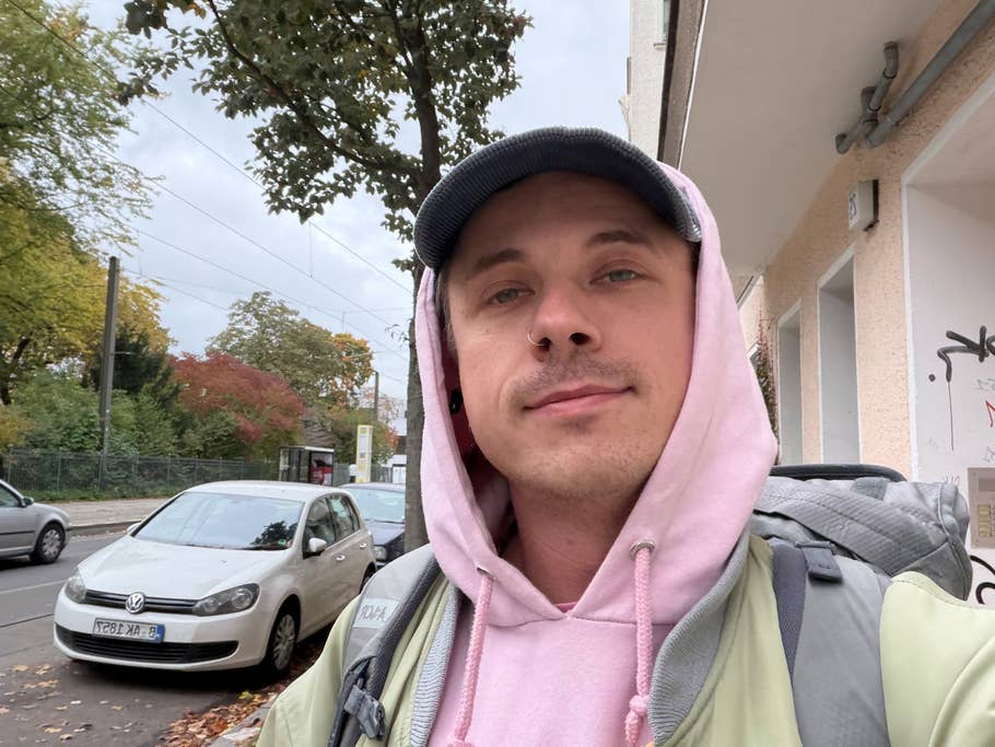 A person wearing a pink hoodie and a cap stands on a street with trees and parked cars.