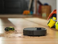 A robotic vacuum cleaner on a wooden floor near scattered soccer balls and shoes.