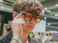 A young man wearing stylish sunglasses, touching his temple, in an indoor event space.