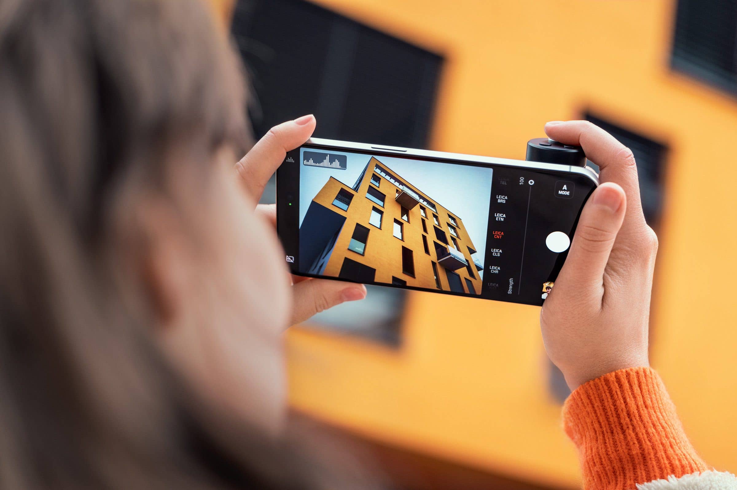 A person holds a Leica LUX Grip camera, capturing a modern yellow building.