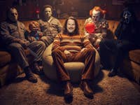 Casi is sitting in an armchair, with numerous horror legends on the couch behind him.