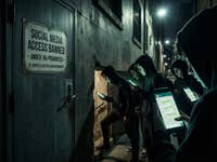A sign announces a social media ban. Children with their eyes on their cell phones and hoodies sneak past the sign into the basement.