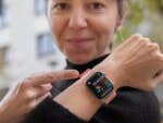 A woman in a black sweater points to her Apple Watch showing time and activity stats.