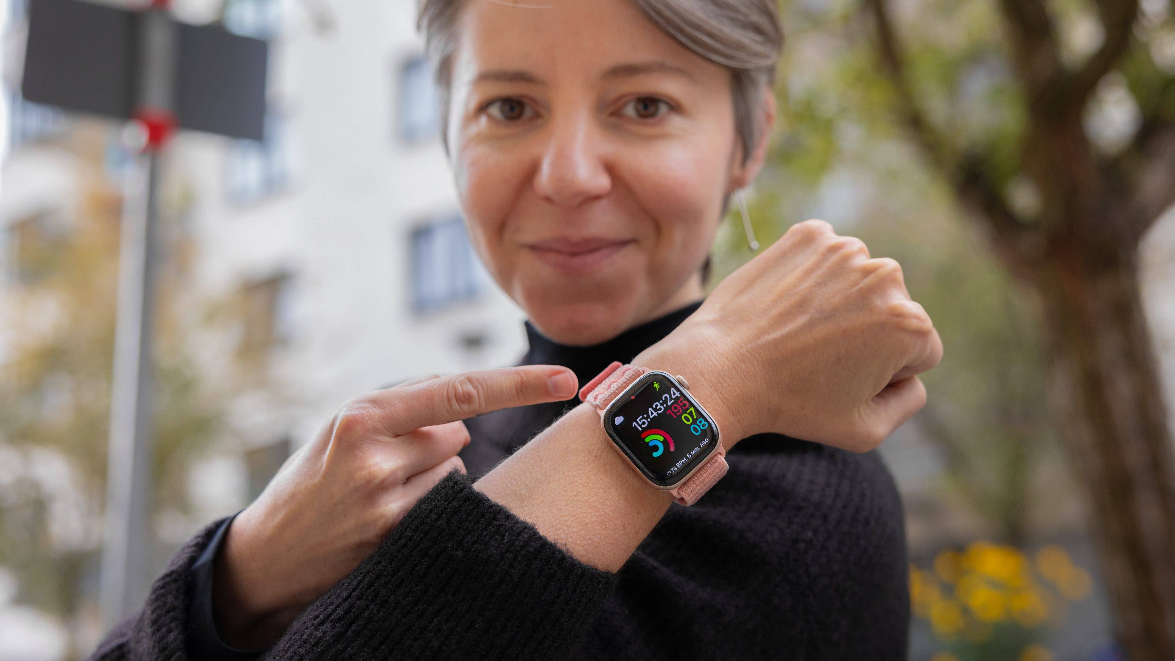 A woman in a black sweater points to her Apple Watch showing time and activity stats.