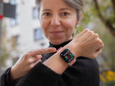 A woman in a black sweater points to her Apple Watch showing time and activity stats.