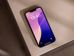 An Apple iPhone 16e lying on a table, displaying the time 13:48 and date February 26.