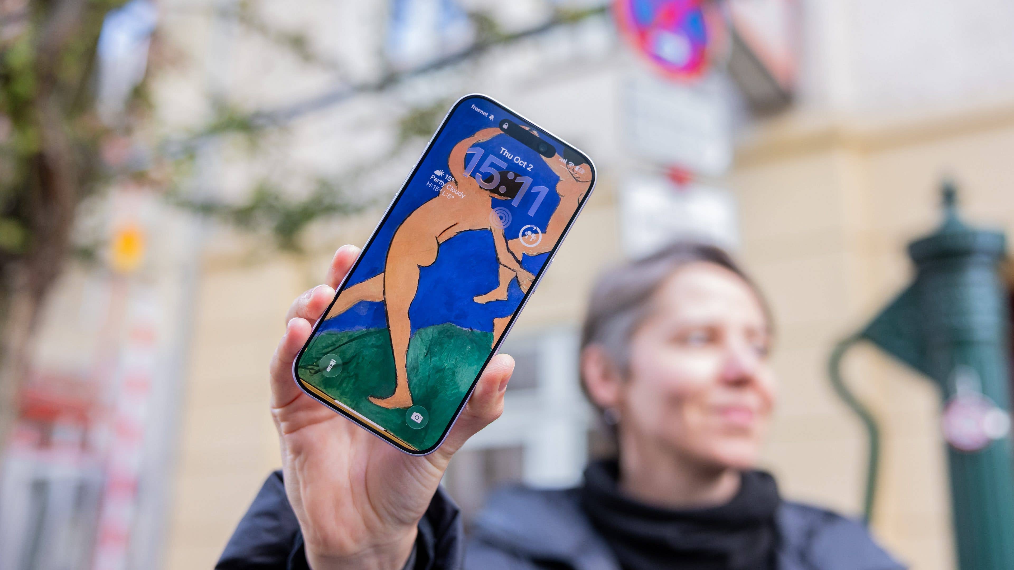 A person holding an iPhone 17 displaying artwork as the lock screen with the time 15:11.