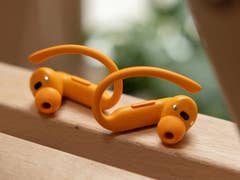 Orange Powerbeats Pro 2 wireless earbuds resting on a wooden surface with blurred greenery in the background.