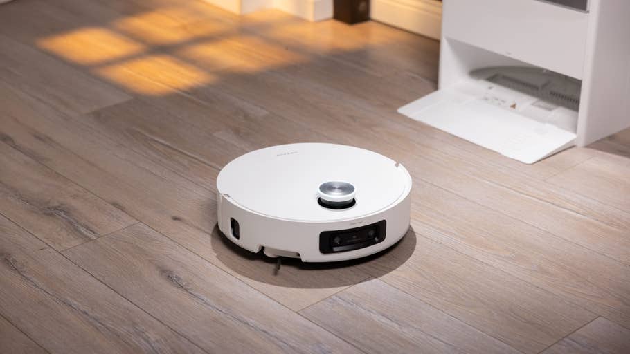 A white robot vacuum cleaner on a wooden floor, with a charging station in the background.