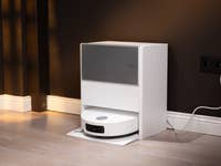 A white automatic vacuum cleaner docked against a dark wall, with a power cord plugged in.
