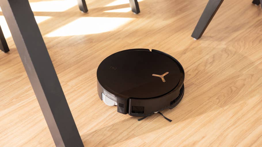 A black Ecovacs Deebot X8 Pro Omni robot vacuum cleaner on a wooden floor, positioned under a chair.