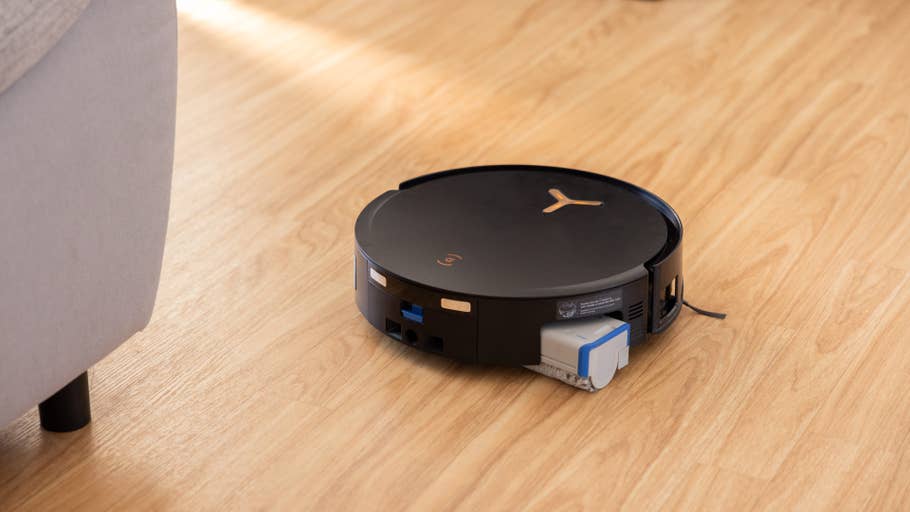 A black Ecovacs Deebot X8 Pro Omni robot vacuum cleaner on a wooden floor next to a sofa.