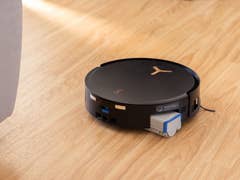 A black Ecovacs Deebot X8 Pro Omni robot vacuum cleaner on a wooden floor near a couch.