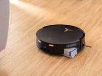 A black Ecovacs Deebot X8 Pro Omni robot vacuum cleaner on a wooden floor near a couch.