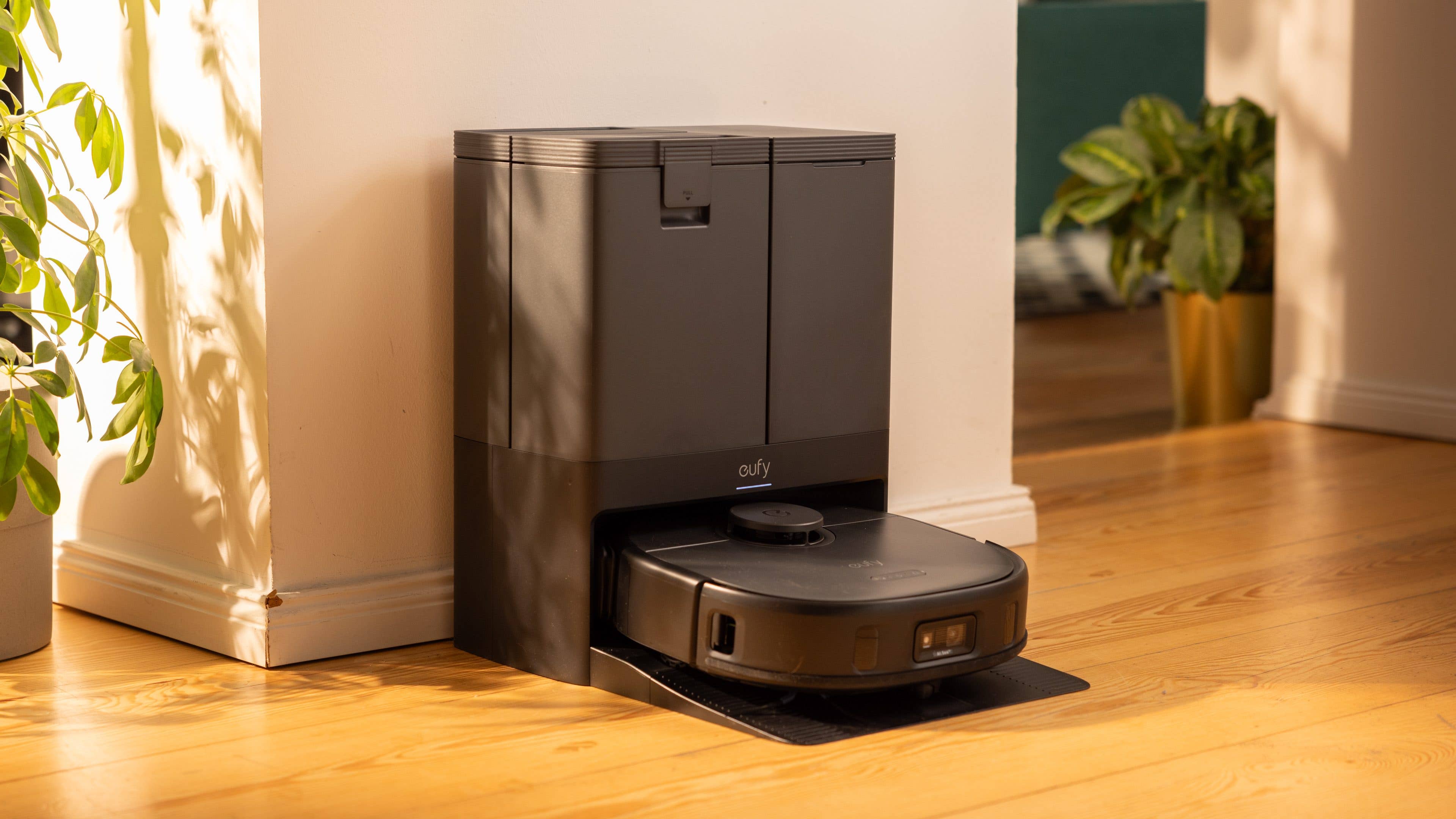 A Eufy X10 Pro Omni robotic vacuum docked next to a plant in a sunlit room.