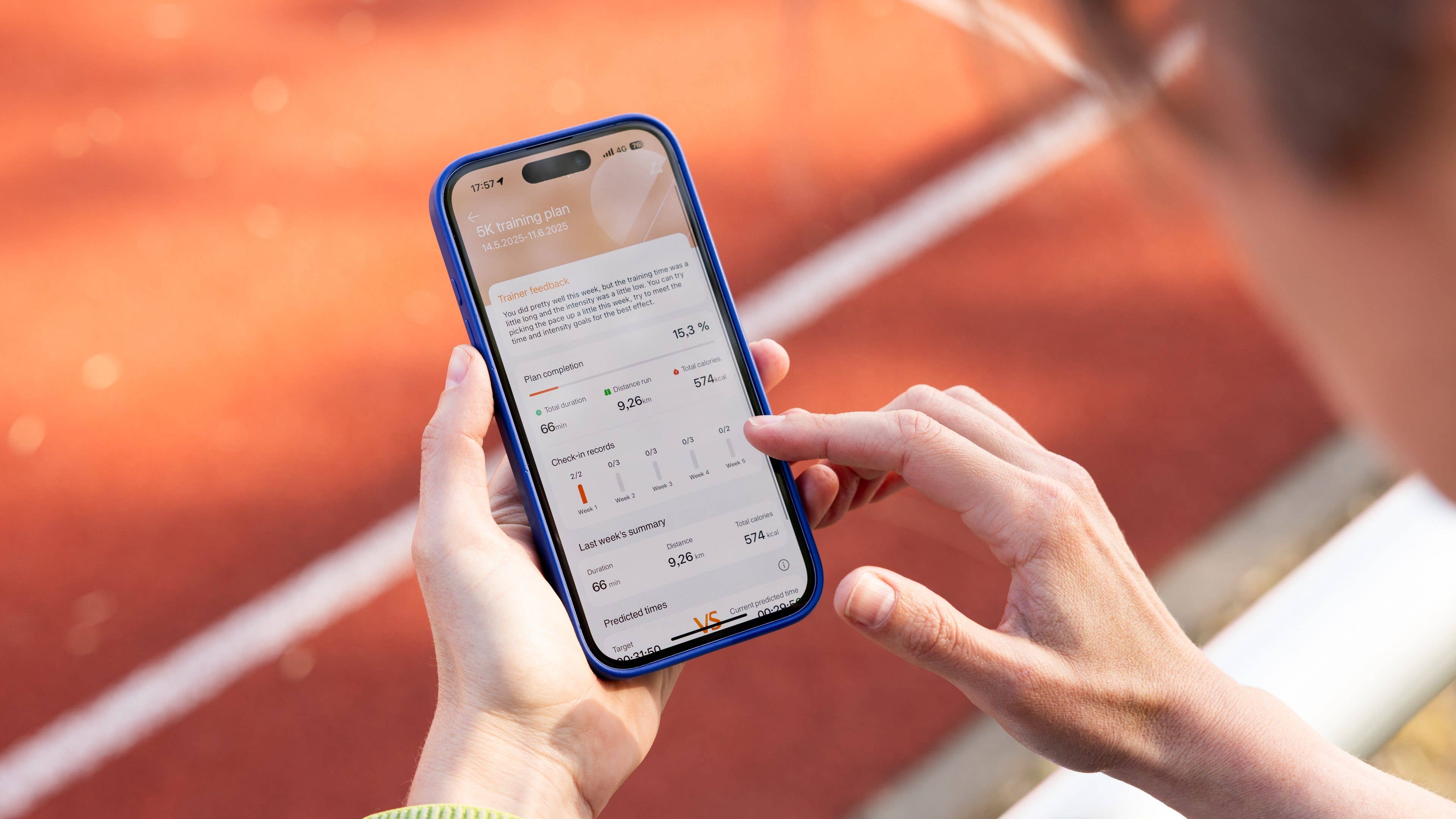 A person holds a smartphone displaying a 5K training plan with performance metrics on a running track.