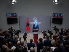 A press conference in Albania with a speaker at a podium, flanked by cameras and an Albanian flag.