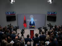 A press conference in Albania with a speaker at a podium, flanked by cameras and an Albanian flag.