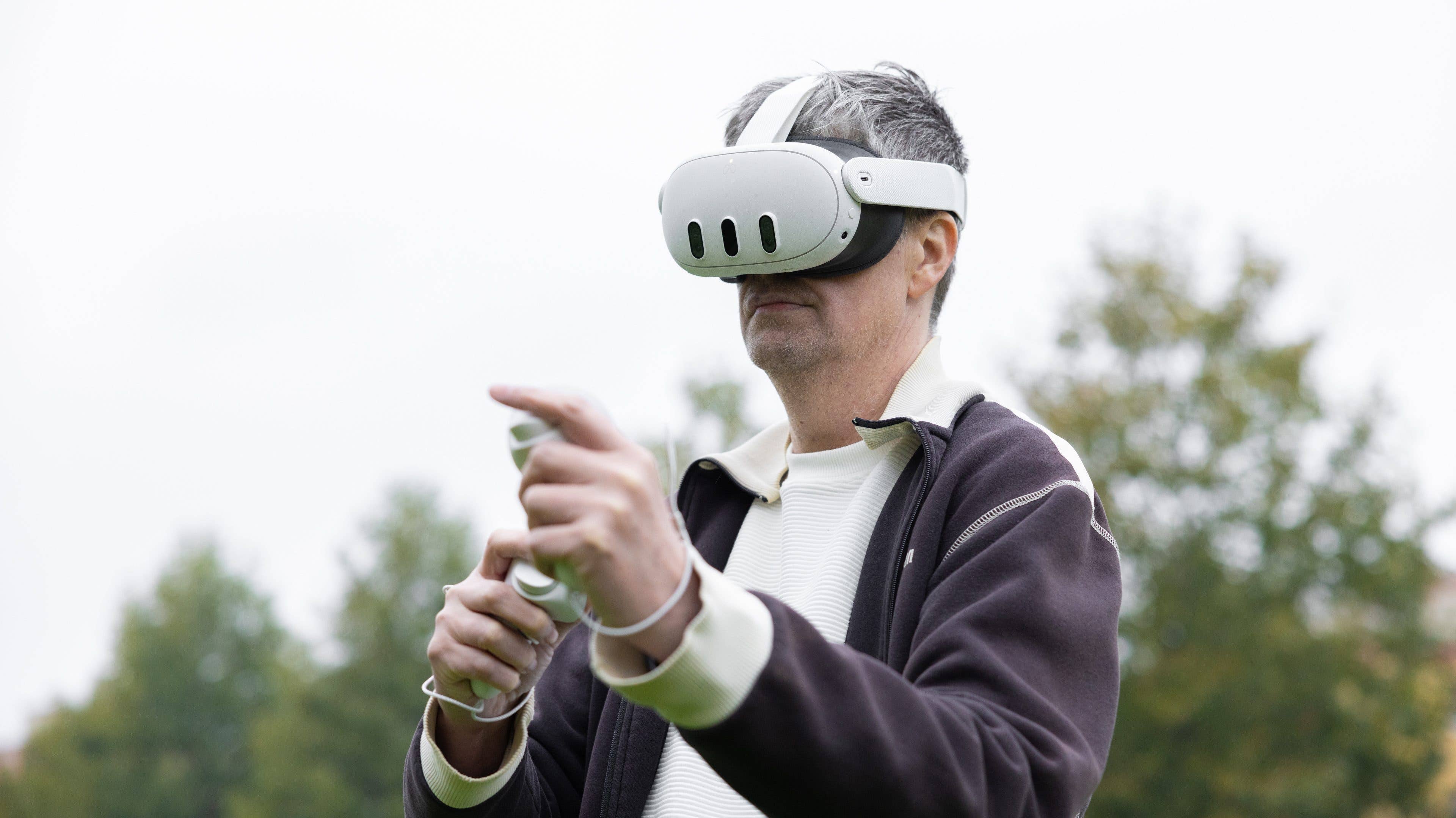 A man wearing a virtual reality headset outdoors, holding a controller and pointing.