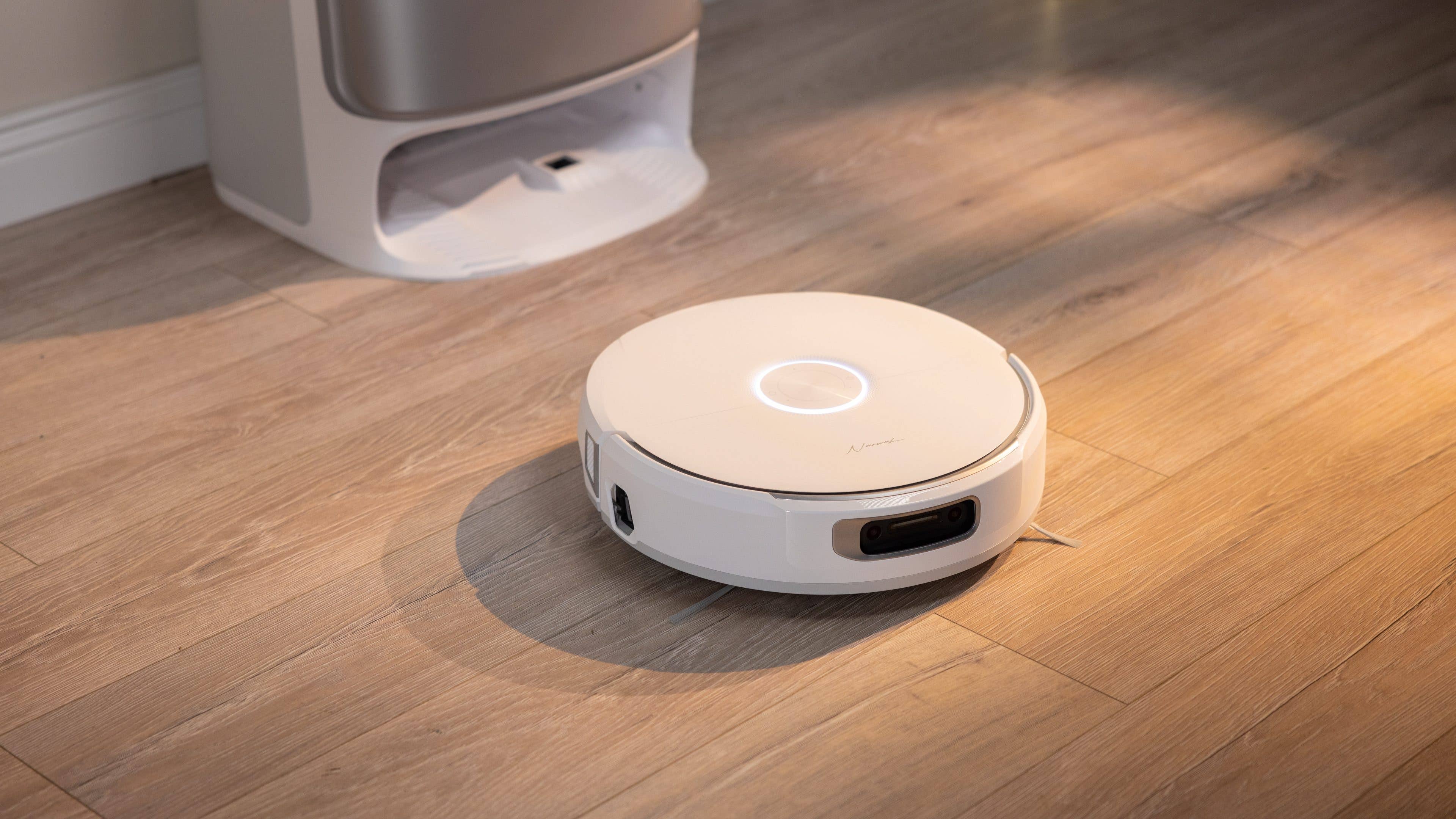 A white robotic vacuum cleaner on wooden floor, with a silver device in the background.