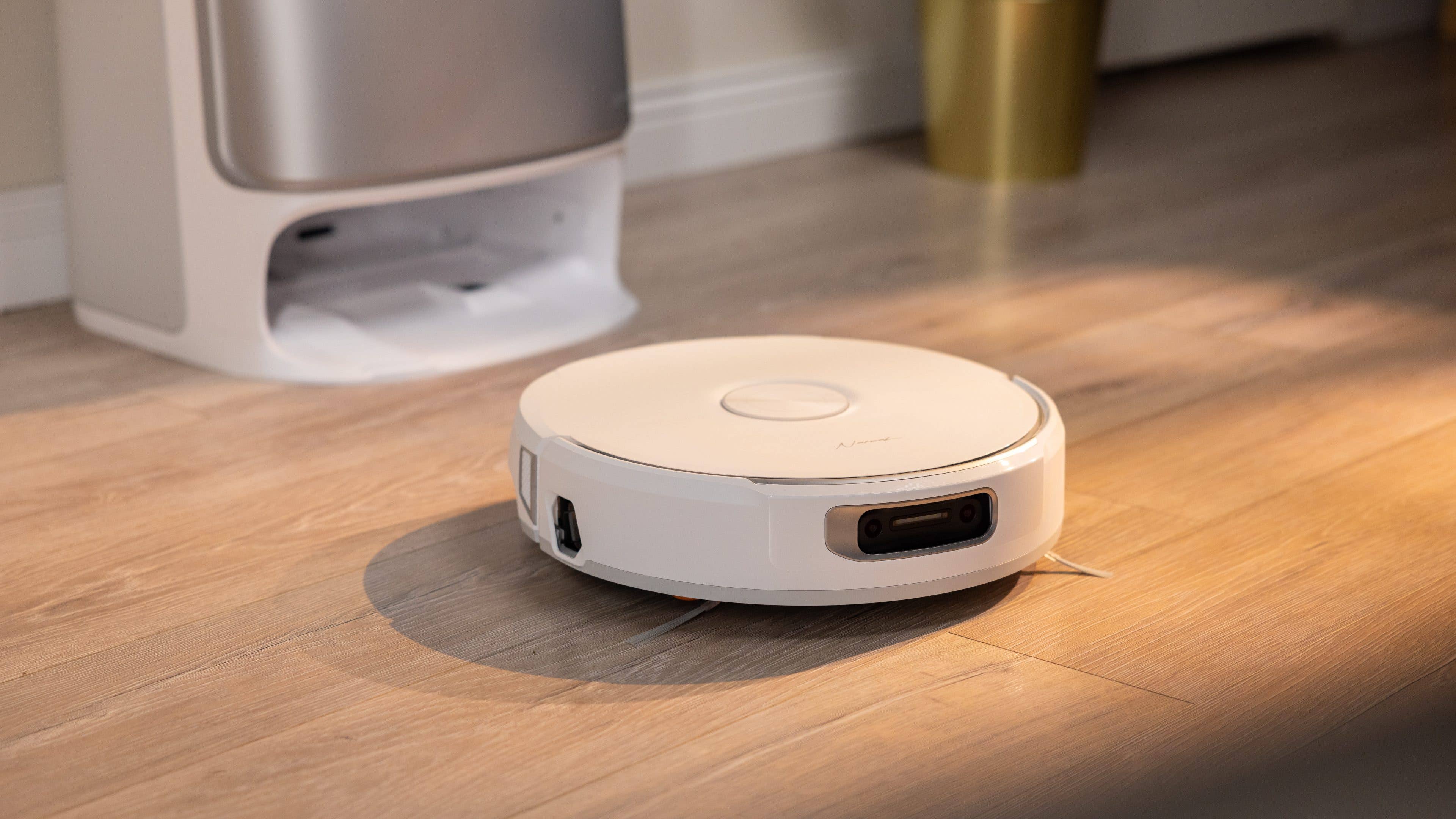 A white robotic vacuum cleaner on a wooden floor near a trash can.