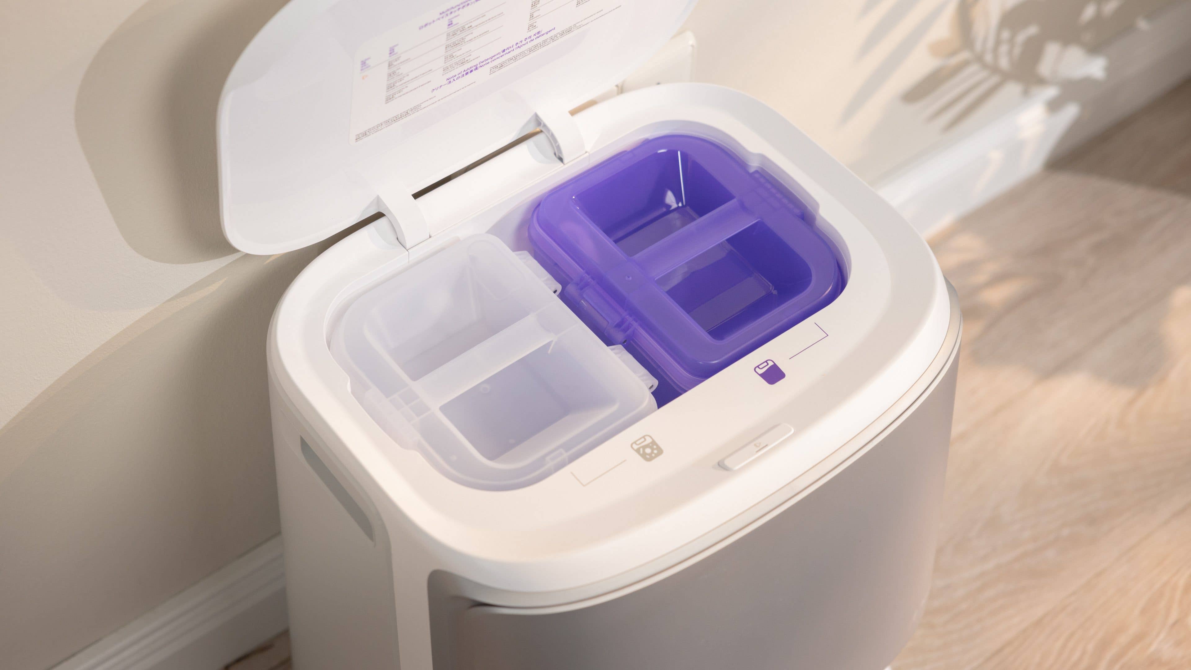 A modern trash bin with two compartments, one purple and one clear, open lid, located against a wall.