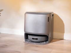 A sleek gray robotic vacuum cleaner sits against a wall in a well-lit room.