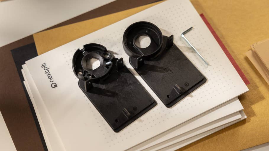Two black plastic parts for a Nuki Smart Lock Pro on a stack of paper, next to an L-shaped tool.