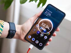 A person holding a OnePlus 13R smartphone displaying the time and battery status, plugged into a charger.