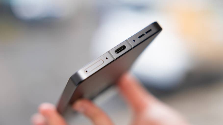 A hand holding the top edge of a OnePlus 13R smartphone, showing the sim card slot and USB-C port.