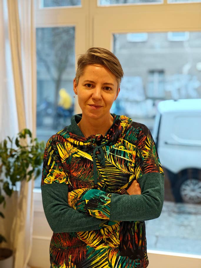 A person with short hair wearing a colorful hoodie stands with arms crossed in a bright room.