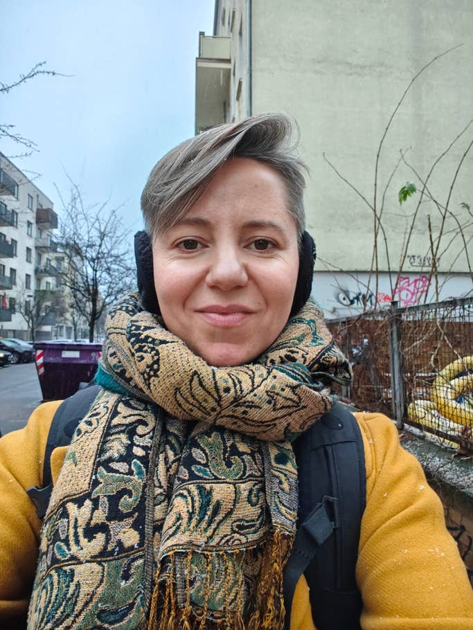 A person with short hair wearing a patterned scarf and headphones, standing outdoors in a city.