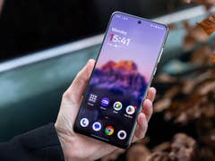 A person holding a OnePlus 13 smartphone displaying the home screen with time and apps.