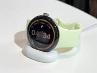 A Google Pixel Watch 4 on a charging dock, displaying time 04:34 and a battery status of 100%.