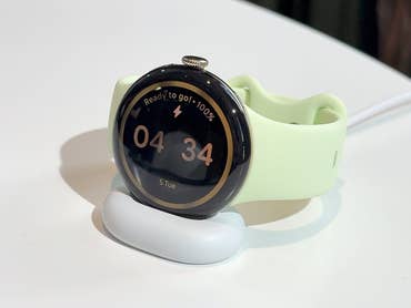 A Google Pixel Watch 4 on a charging dock, displaying time 04:34 and a battery status of 100%.