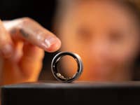 A close-up of a Samsung Galaxy Ring on a stand, with a hand reaching to touch it in the background.