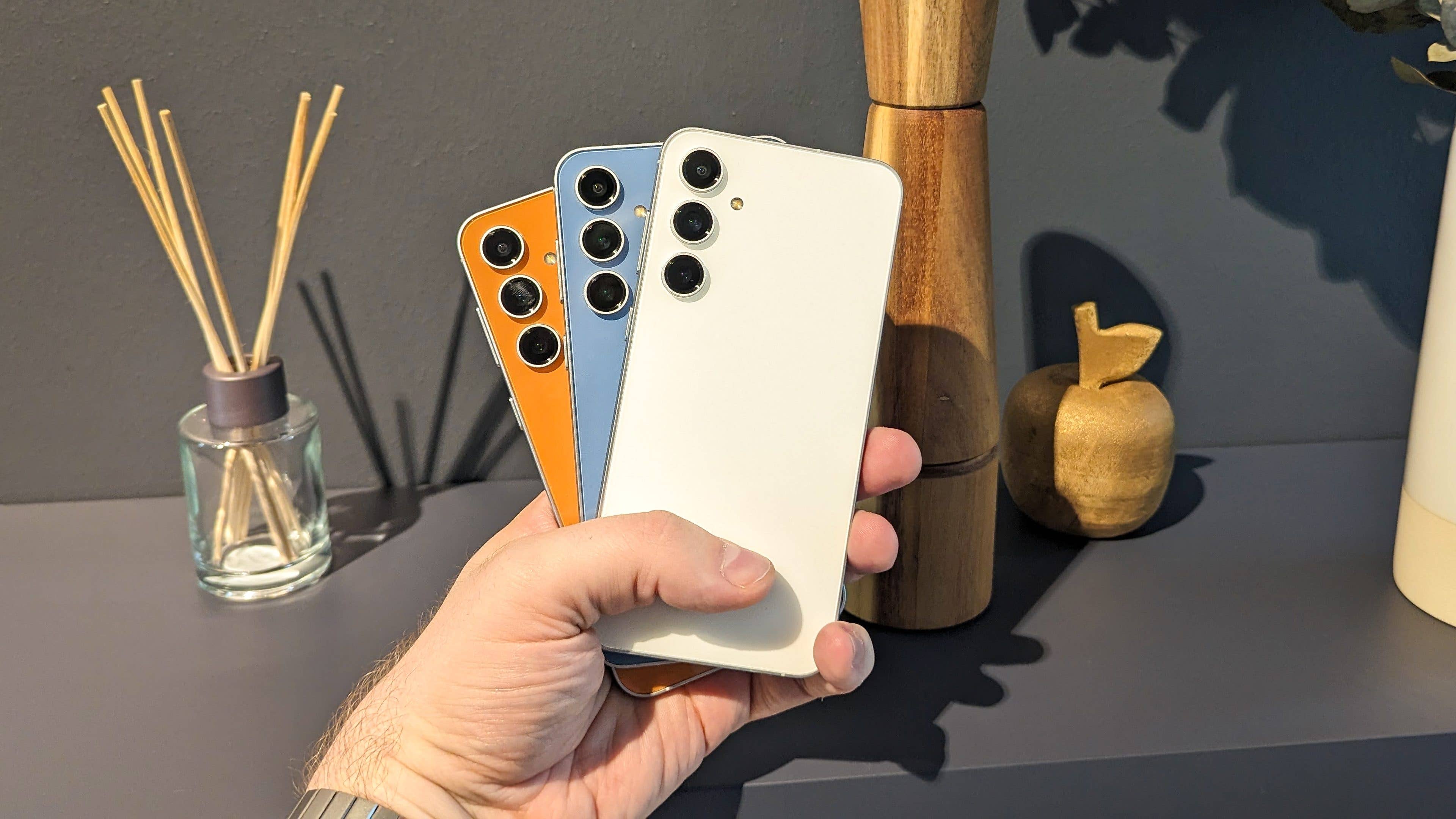 Three different colored models of the S23 FE held in front of a kitchen shelf in hand Three different colored models of the S23 FE held in front of a kitchen shelf in hand