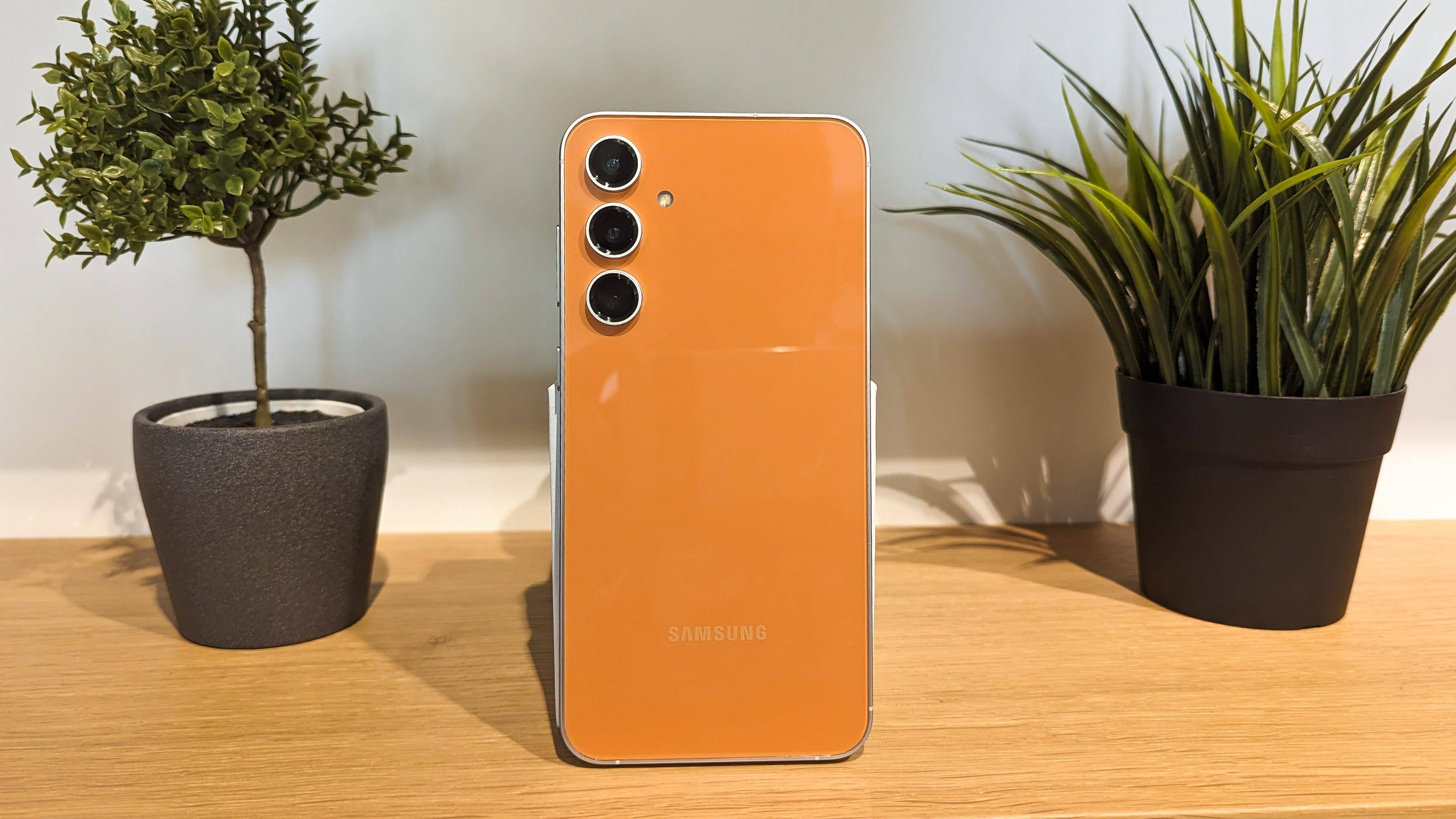Galaxy S23 FE in tangerine color stands between two houseplants on a shelf