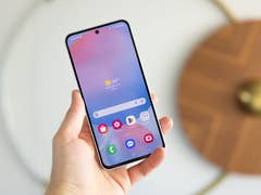 A person holding a Samsung Galaxy S24 FE smartphone displaying a weather app with a sunny 34°C.