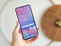 A person holding a Samsung Galaxy S24 FE smartphone displaying a weather app with a sunny 34°C.