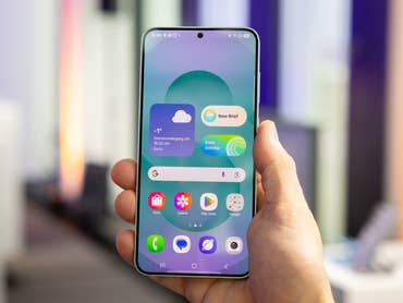 A person holding a Samsung Galaxy S25 smartphone displaying a weather widget and app icons.