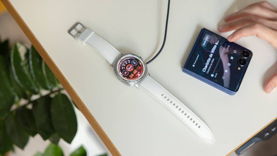 Samsung Galaxy Watch 8 Classic on a table, charging, next to a smartphone playing music.