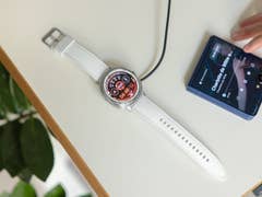 Samsung Galaxy Watch 8 Classic on a table, charging, next to a smartphone playing music.