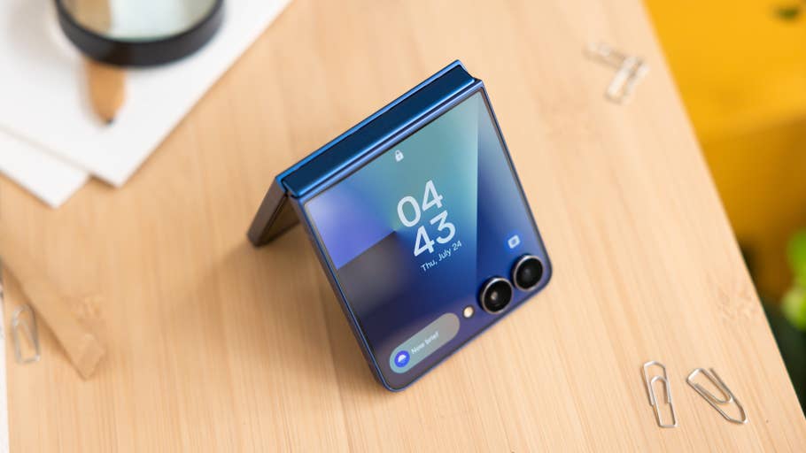 A blue Samsung Galaxy Z Flip7 smartphone displayed on a wooden surface showing the time 04:43.