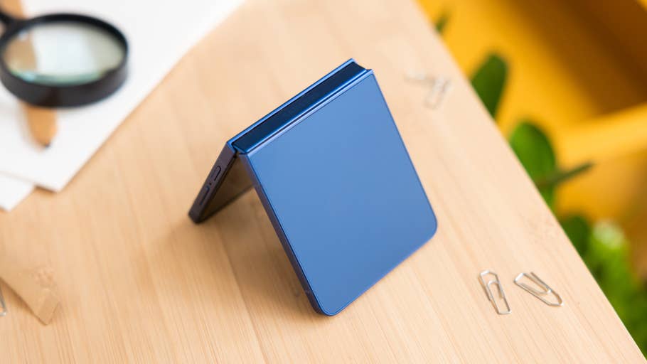 A Samsung Galaxy Z Flip7 smartphone in blue, partially open on a wooden surface with office supplies nearby.