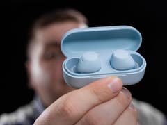 A person holding a blue charging case with wireless earbuds inside, against a dark background.