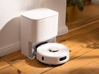 A white robotic vacuum cleaner positioned next to a charging station on wooden flooring.