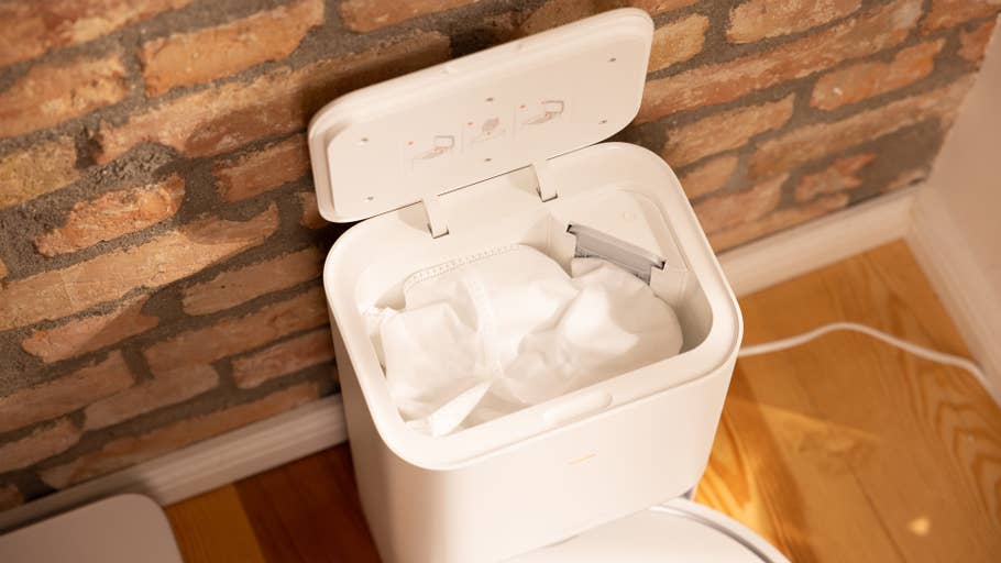 A white smart trash can with an open lid showing a bag and a clean interior in front of a brick wall